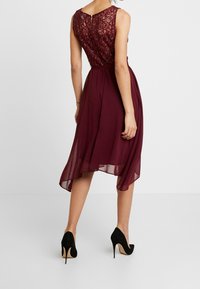 Kvinna klädd i en ärmlös bourgogne laceklänning med asymmetrisk fåll och svarta högklackade skor, stående mot en enkel vit bakgrund.