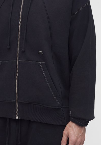 Κοντινό πλάνο ατόμου που φοράει μαύρο φούτερ με φερμουάρ, λευκές ραφές, εμπρόσθιες τσέπες και μικρό λογότυπο "ACW" στο στήθος.