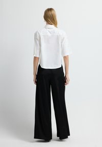 Camisa blanca de manga corta con patrones decorativos y un dobladillo que termina por encima de unos pantalones de pierna ancha negros. La parte trasera presenta un diseño liso.