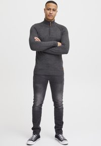 Maglione grigio con zip al collo, dettagli a coste su polsini e orlo, abbinato a jeans scuri e sneaker nere alte, in piedi con le braccia incrociate.