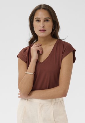 Femme aux cheveux bruns portant un chemisier marron à col en V, un pantalon crème, un bracelet argenté et des boucles d'oreilles, posant avec la main près du visage sur fond blanc.