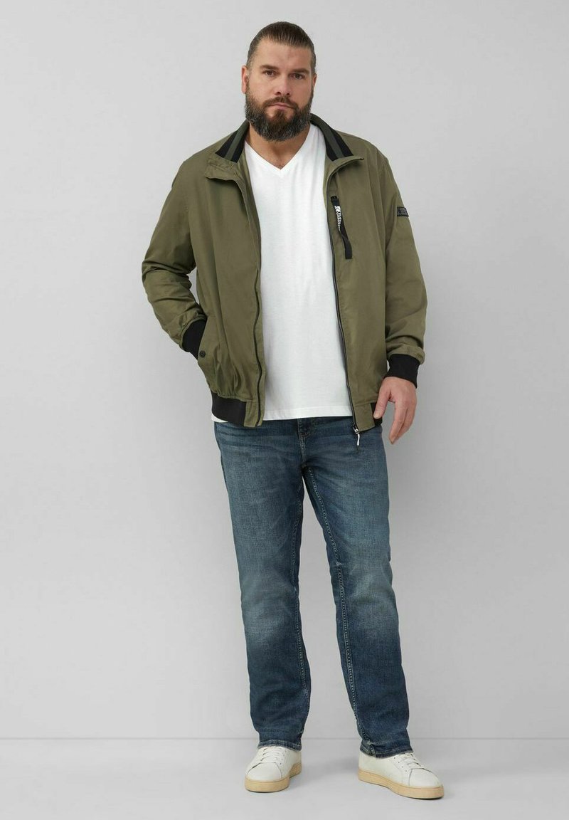 Olivgrüne Bomberjacke mit schwarzem Kragen, Reißverschluss vorne und gerippten Bündchen. Getragen über einem weißen V-Ausschnitt-T-Shirt und blauen Jeans. Weiße Sneaker.