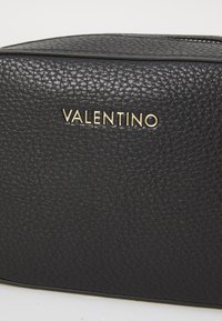 Borsa in pelle nera con una texture a granuli, caratterizzata da un logo "VALENTINO" in oro. Il design include una chiusura con zip lungo la parte superiore.