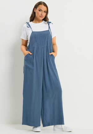 Kvinde iført blå buksedragt med brede ben og bindebånd på skuldrene samt hvid T-shirt med korte ærmer, hænderne i lommerne, stående mod en lys baggrund.