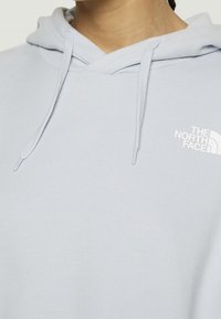 Sweat à capuche bleu clair avec cordon de serrage, arborant un logo blanc sur la poitrine. Le matériau semble doux et texturé, avec une coupe décontractée.