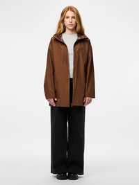 Καφέ αδιάβροχο μπουφάν με κουκούλα, σχεδιασμένο με μπροστινό φερμουάρ και ελαφρύ υλικό. Συνδυάζεται με μαύρο παντελόνι φαρδιάς γραμμής και μαύρα παπούτσια.