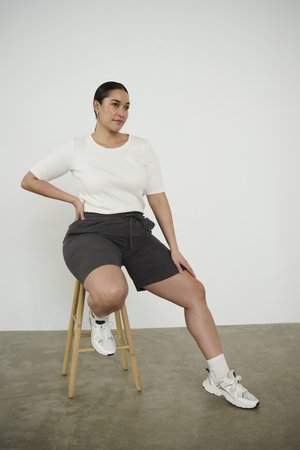 Kvinde med mørkt hår iført en hvid skjorte og grå shorts, der sidder på en træskammel mod en ensfarvet hvid væg.