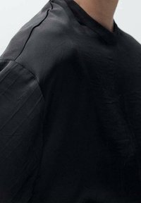 Haut noir à manches courtes en tissu lisse, avec un col rond et une coupe décontractée, présentant une texture subtile et des détails minimalistes.