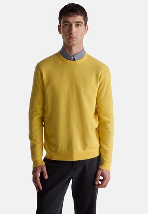 Gelber Strickpullover mit Rundhalsausschnitt und gerippten Bündchen, kombiniert mit einem karierten Hemd darunter und einer schwarzen Jeans.