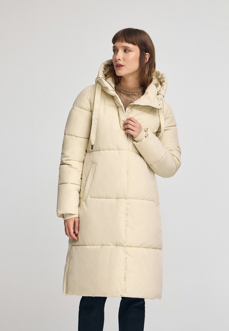 Beżowy ocieplany zimowy płaszcz z kapturem, z frontowym zamkiem błyskawicznym, bocznymi kieszeniami i pikowanym wzorem, sięgający do połowy łydki.