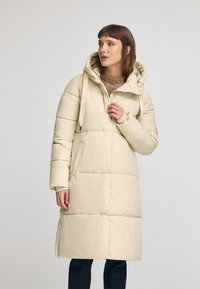 Cappotto invernale imbottito beige con cappuccio, dotato di una chiusura frontale con zip, tasche laterali e un design trapuntato, che arriva fino a metà polpaccio.