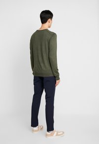 Suéter verde-oliva com decote redondo, textura de malha fina. Combinado com calças azul-marinho e ténis com detalhes beige. Vista traseira apresentada.