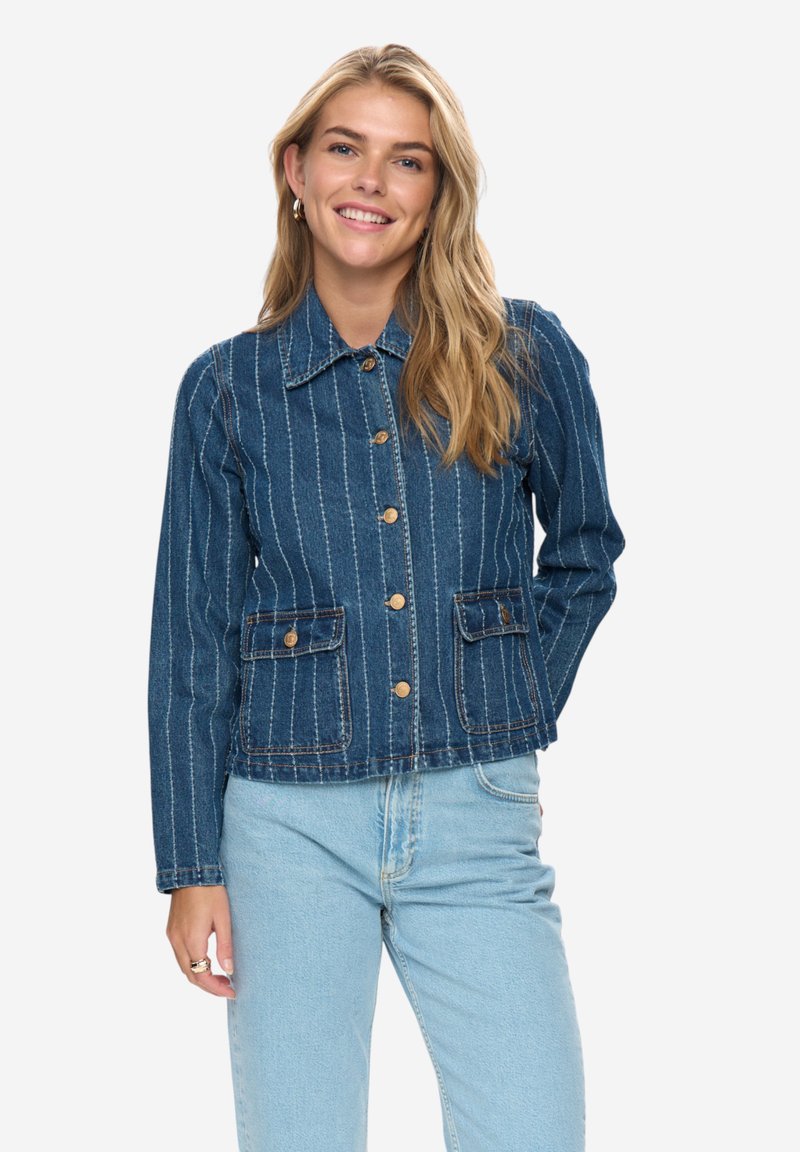 Femme souriante aux longs cheveux blonds portant une veste en denim bleu rayée avec des boutons en laiton et un jean bleu clair sur un fond uni.