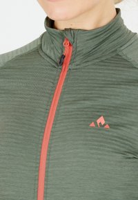Grüner sportlicher Zip-Up-Oberteil mit hohem Kragen, orangefarbigem Reißverschluss, strukturiertem Stoff und kleinem Logo auf der linken Seite.