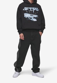 Svart huvtröja med grafisk design och texten "Speed of Life". Matchas med lösa grå jeans och vita sneakers. Enkel, avslappnad stil.