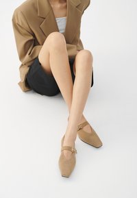 Beige sukové boty s špičatou špičkou, kotníkovým páskem se zlatou sponou a elegantním designem, v kombinaci s blejzrem a sukní.