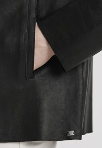 Schwarze Lederjacke mit einem eleganten Design, glatter Textur, seitlichen Taschen und einem metallischen Markenschild nahe dem Saum.