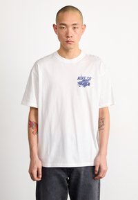 Bílý bavlněný tričko s krátkým rukávem, s modrou grafickou potiskem auta a textem "NIKE SB" na levé straně hrudi.