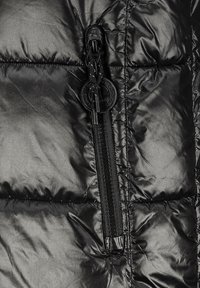 Gros plan sur un tissu de veste matelassée noire brillante featuring une poche zippée verticale avec un tirette de fermeture éclair rond avec logo.