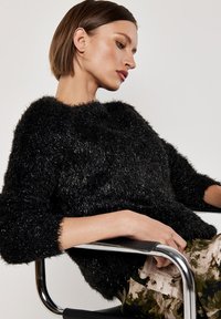 Schwarzer, flauschiger Pullover mit Rundhalsausschnitt und Dreiviertelärmeln; getragen über gemusterten Hosen, kombiniert mit einem chromfarbigen Stuhl.