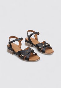 Sandales en cuir noir avec une bride au niveau de la cheville, des accents cloutés et un petit talon. Le design comprend des brides entrecroisées et une semelle marron clair.