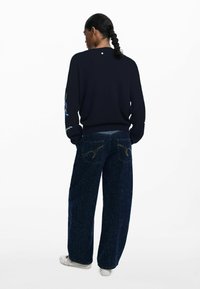 Navy knitted sweater with long sleeves, featuring colorful embroidery on arms. Paired with wide-leg dark blue jeans with distinct back pockets.