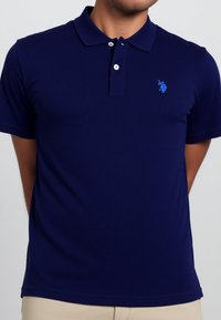 A person wears a navy blue polo shirt with a small blue logo on the left chest, standing with hands clasped behind.