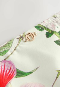 Bloemig bedrukte stof met roze en rode bloemen op een lichte achtergrond, voorzien van een gouden ronde knoop en witte draaddetails.