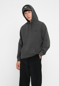 Hanorac gri închis cu buzunar frontal tip kangaroo, glugă cu șnur și un logo mic pe piept. Asortat cu pantaloni din corduroy negri.