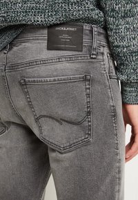 Detailní záběr osoby v šedých denimových džínách s černou nášivkou Jack & Jones nad zadní kapsou a v zeleném pleteném svetru.