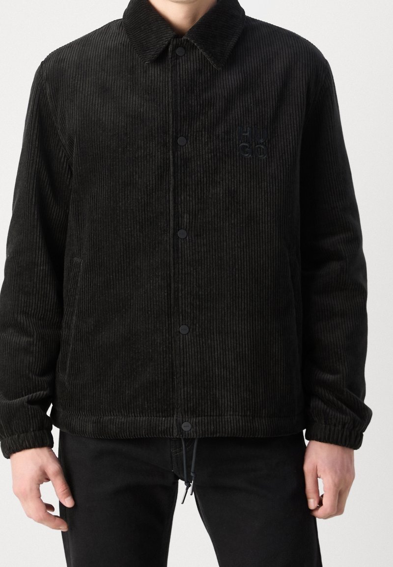 Zwarte corduroy jas met een knoopsluiting aan de voorkant, kraag en elastische manchetten. Beschikt over een logo op de borst en zijzakken. Zachte textuur.