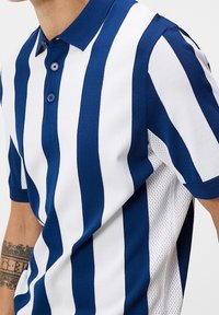 Polo tričko s modro-bílými vertikálními pruhy, vybavené dvěma knoflíky, límečkem a texturovanou boční stranou pro prodyšnost.
