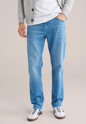 Man draagt lichtblauwe spijkerbroek, witte sneakers met zwarte strepen, grijze cardigan en wit shirt, staat met één hand in de zak.