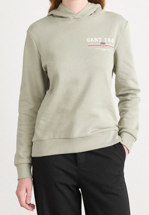 Personne portant un sweat à capuche vert clair avec un logo GANT 1949 sur la poitrine et un pantalon noir, une main dans la poche.