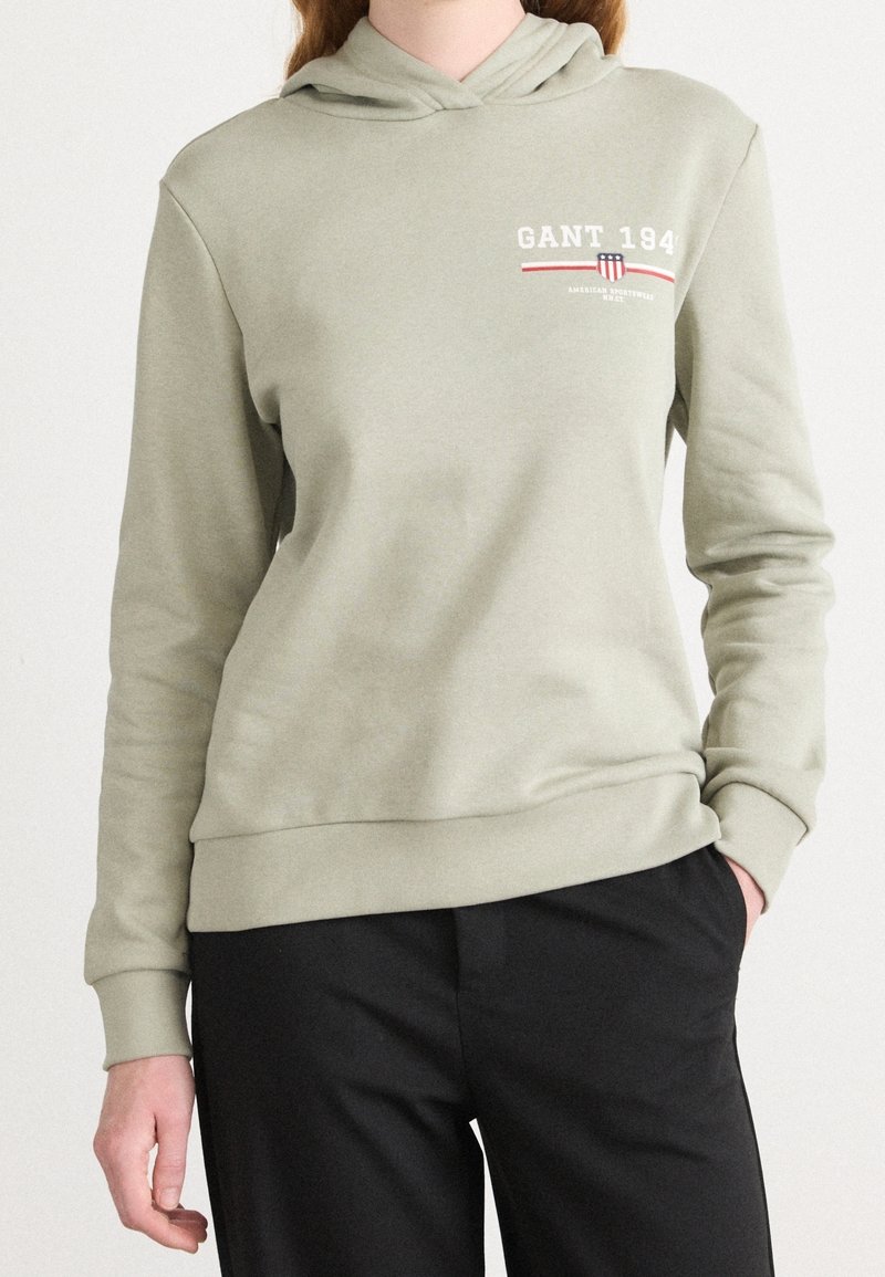 Personne portant un sweat à capuche vert clair avec un logo GANT 1949 sur la poitrine et un pantalon noir, une main dans la poche.