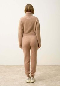 Beżowy sweter dzianinowy z wysokim kołnierzem i długimi rękawami, w komplecie z dopasowanymi spodniami, oba wykonane z miękkiego, teksturowanego materiału. Casualowe obuwie.