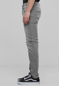 Harmaat slim-fit farkut, joissa on kulutusta nivelissä, yhdistettyinä mustiin matalavartisiin lenkkareihin, joissa on valkoisia korostuksia ja nauhoja.