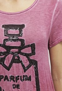 Růžové tričko s krátkým rukávem, zdobené černým flitrovým potiskem parfémové láhve s mašlí a textem "PARFUM DE."