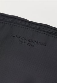 Schwarze Reißverschlusstasche mit strukturiertem Oberflächendesign. Mit geprägtem Branding "ARKK COPENHAGEN EST. 2015" auf dem Stoff. Minimalistisches Design.