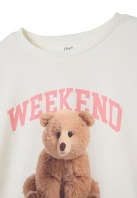T-shirt blanc avec un motif d'un ours en peluche marron, accompagné du texte rose "WEEKEND" au-dessus de l'image de l'ours. Matière en coton doux.