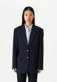 Blazer bleu marine avec épaules structurées, deux poches avant, boutons en métal doré et une texture de tissu lisse, porté sur une chemise rayée.