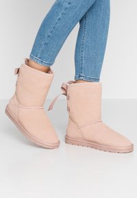 Botas de tobillo de ante rosa con un interior suave, punta redonda y detalle de lazo en el costado. Suela de goma plana, tacón de una pulgada.