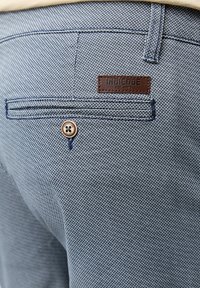 Blue textured fabric of trousers with a brown leather label marked "INDICODE" and a button detail, featuring a back pocket design.