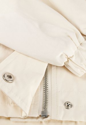 Close-up van beige stof met een deels open ritssluiting met drukknoopjes aan beide zijden en een elastische manchet van een mouw.