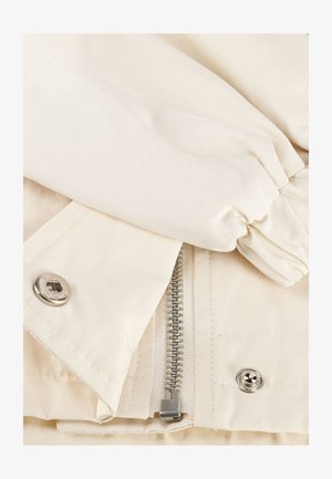 Close-up van beige stof met een deels open ritssluiting met drukknoopjes aan beide zijden en een elastische manchet van een mouw.