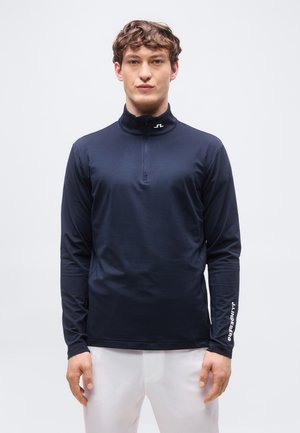 Vyras dėvi tamsiai mėlyną ilgomis rankovėmis užsegamą megztinį su aukštu apykakliu, baltus švarko „J.LINDEBERG“ užrašus ant rankovės ir baltas kelnes, stovintis prieš paprastą foną.