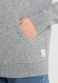 Pull gris en maille avec une poche latérale, des poignets et un ourlet côtelés. Le tissu a une texture douce avec une étiquette logo sur le côté inférieur.