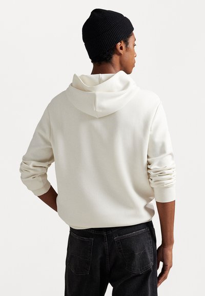 C hoodie branco com uma textura suave, apresentando um capuz com cordão e mangas dobradas, combinado com jeans escuros.