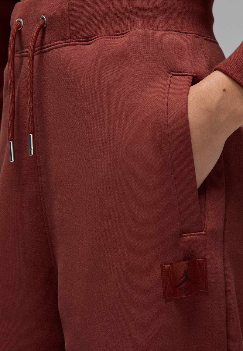 Bordeauxfarbene Sweatpants mit einer glatten Textur, ausgestattet mit einem Logo-Patch und einem Kordelzug mit metallenen Enden. Zwei in das Design integrierte Seitentaschen.