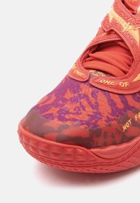 Czerwone i fioletowe buty sportowe z teksturowaną cholewką, unikalnym wzorem w kamuflaż i wyraźnymi pomarańczowymi akcentami na języku i bokach.